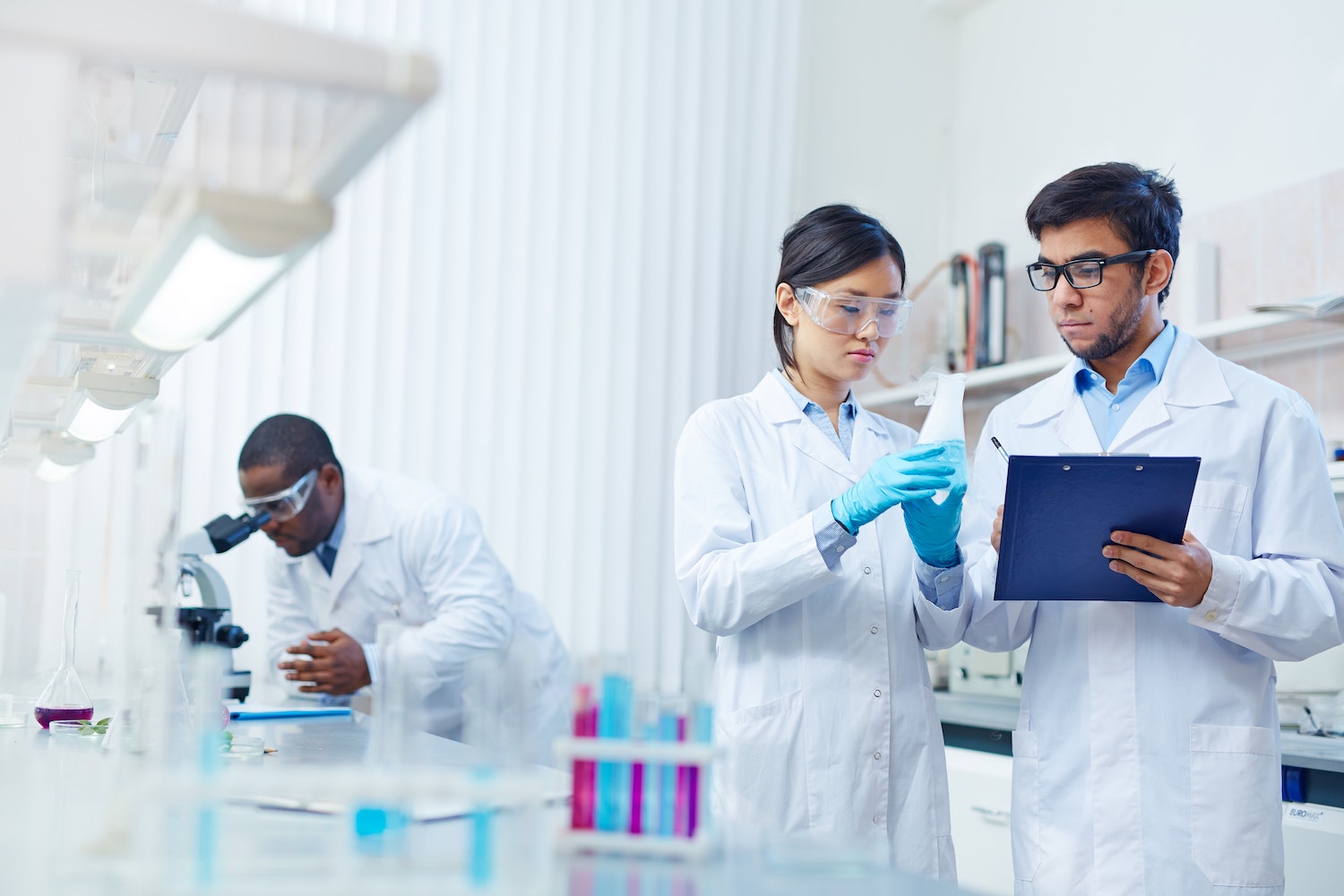 man and woman scientists in lab coats look into a beaker as another male scientist looks into a microscope
